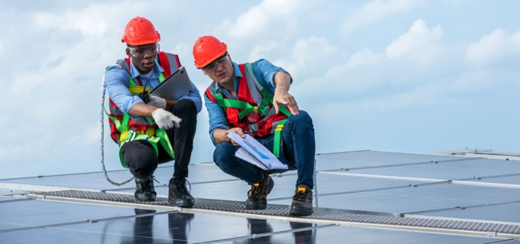 Two men in safety gear examine a solar panel on a commercial roof, highlighting financing programs for roof replacement.