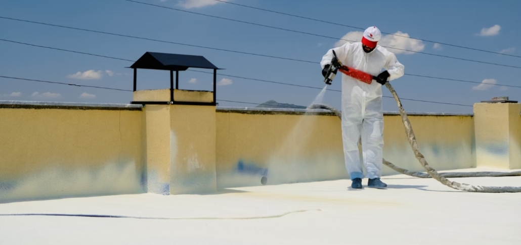 A man in a white suit is spraying white paint on a flat roof, illustrating the process of applying spray polyurethane foam.