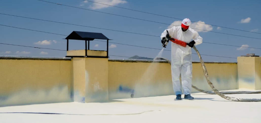 A man in a white suit applies white spray paint to a roof, demonstrating spray foam roofing techniques.