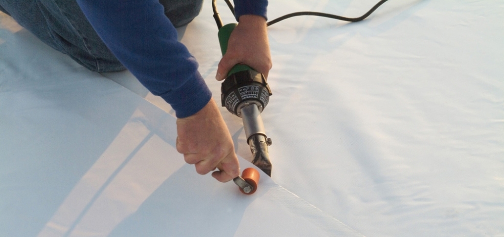 A person employs a power heat and roller to the white fabric, essential for heat-welded seams in TPO roof installation.