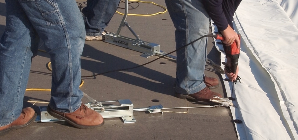 Two men are on a roof, collaborating to secure a large white fabric membrane during a TPO roof installation.