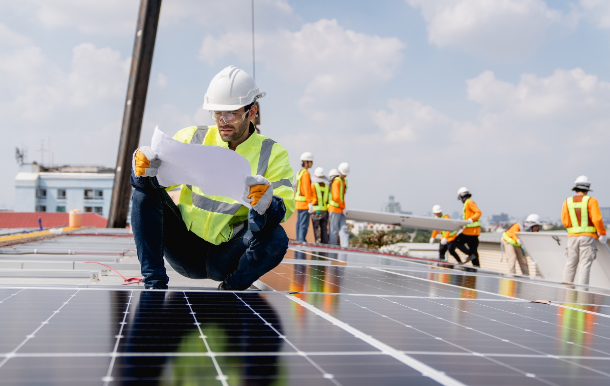 A worker wearing a hard hat and safety vest is installing solar panels on a warehouse roof.