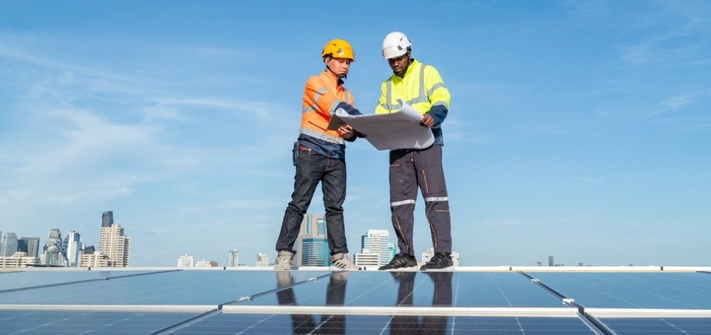 Two men wearing hard hats and safety vests inspect a rooftop with solar panels, focusing on layout for commercial solar use.